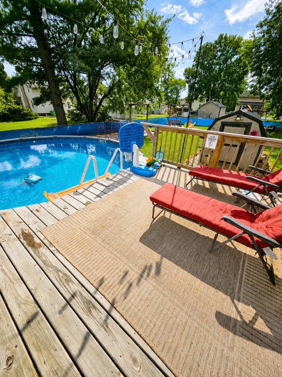 Blue pool beside a wooden deck with two red cushioned loungers, string lights and a pool basketball hoop
